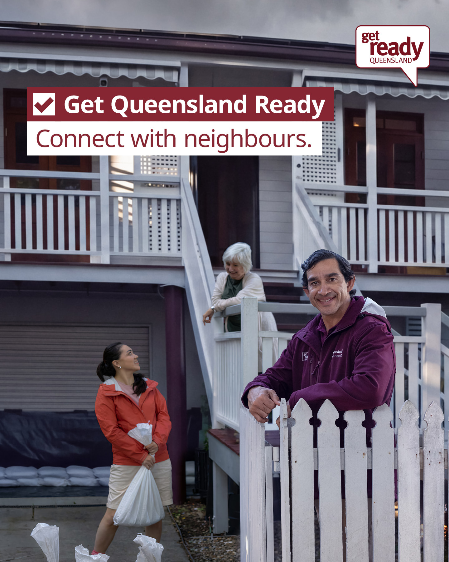 A man leans on a fence smiling whilst an older lady on the stairs behind him looks down at a younger lady lifting a sandbag 