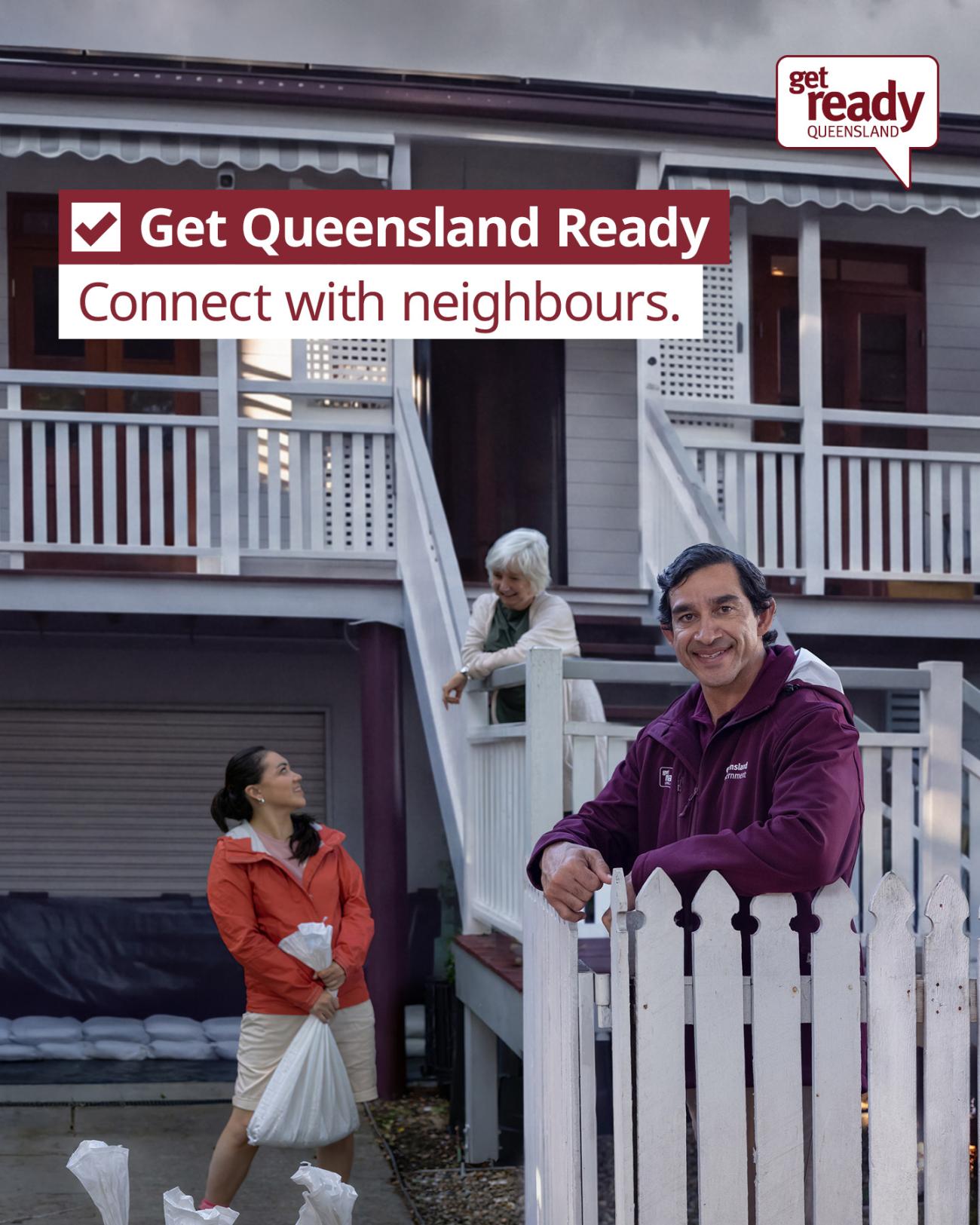 A man leans on a fence smiling whilst an older lady on the stairs behind him looks down at a younger lady lifting a sandbag 