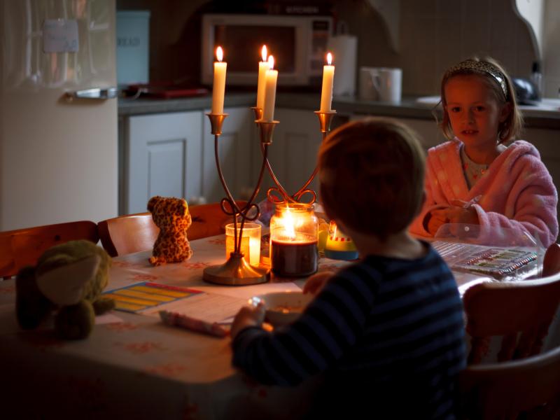 The power has gone out and two children are sitting together at a table at home with candles in front of them for light