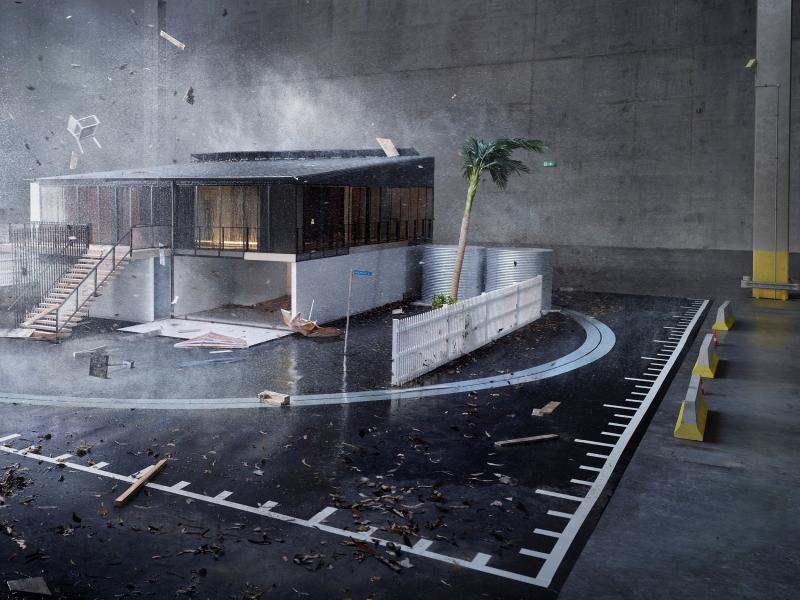 This is a picture of the One House model. The photo shows a classic Queenslander home that is built to withstand cyclonic winds, bushfire and flooding. The home is shown in a warehouse-style testing station where the home is being tested for its resilience to heavy rain, strongs winds and debris. The picture shows strong winds picking up items around the home that become dangerous and damaging in a cyclone including a garden chair, rubbish bins, tree branches, sheets of tin and planks of wood. 